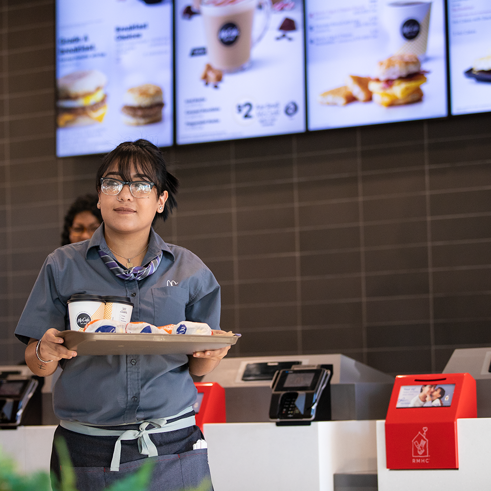 A McDonald's customer carrying their meal on a dine-in tray away from the register, a bright red donation box is featured to the right, breakfast items in soft focus on the menu display