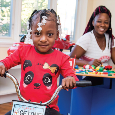 RMHC family, daughter on bike with Alabama RMHC license plate, mom sitting at play table in soft focus background