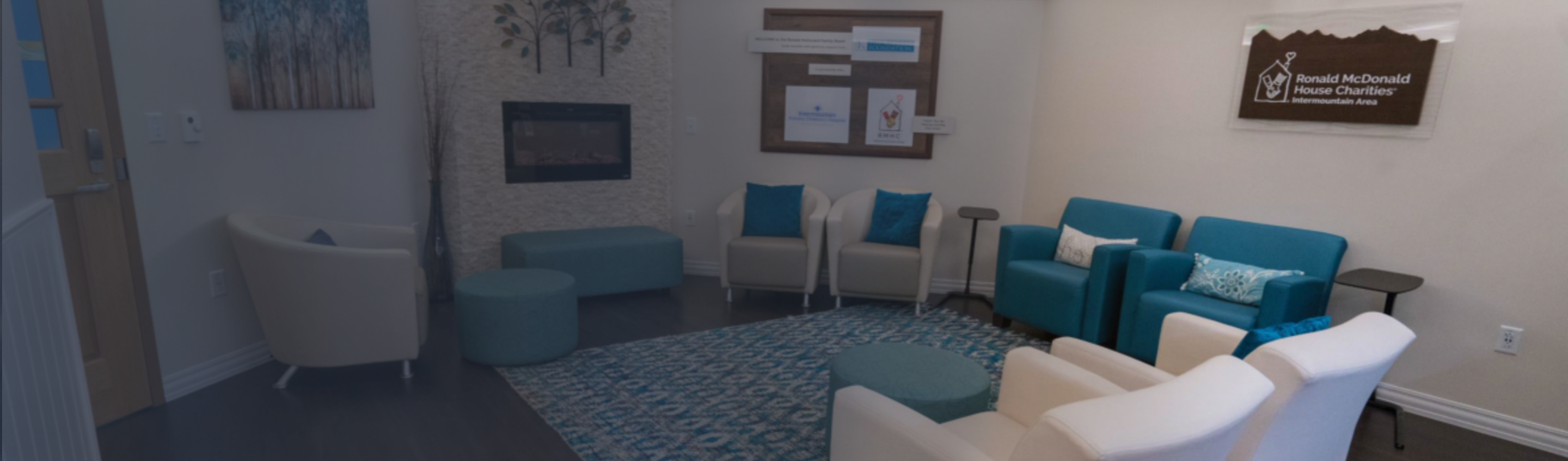 A hospital's Ronald McDonald Family Room sitting room, counter clockwise from the bottom, alternating pairs of white and teal armchairs, a settee and ottoman pair, and a lone white armchair circle 3 sides of a teal, patterned rug