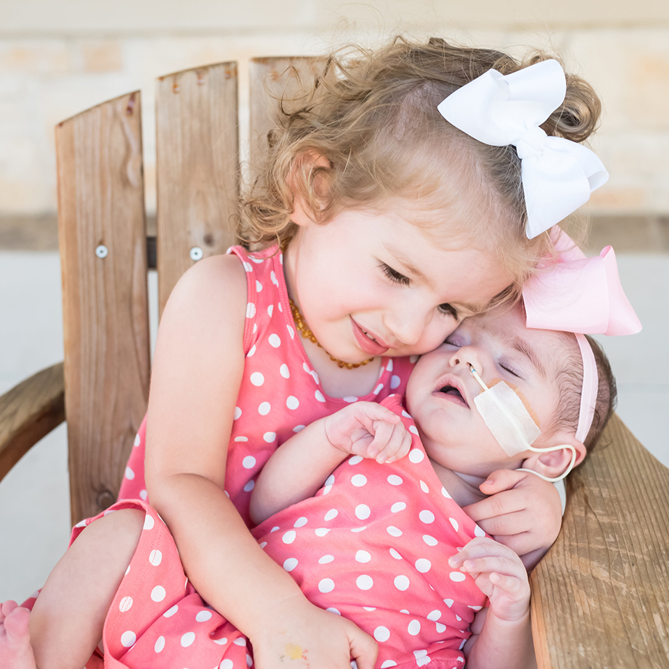 RMHC family in matching polka-dotted, pink dresses, a toddler sitting in a miniature Adirondack chair holding infant sibling in treatment